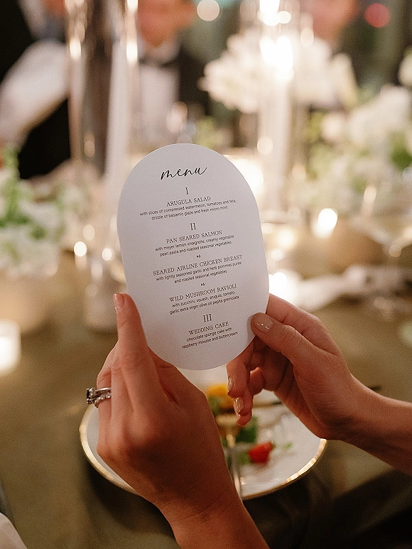 Wedding menu card with arched wedding menu typography beside a ring, candlelight, and greenery on a reception table with warm lights