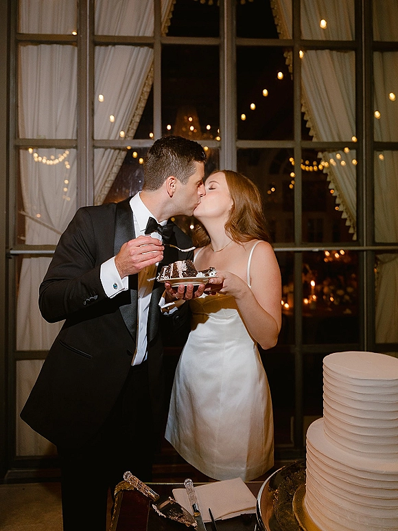 Wedding cake cutting as bride and groom kiss beside a slice on dessert plates, with string lights and window panes in a nighttime reception