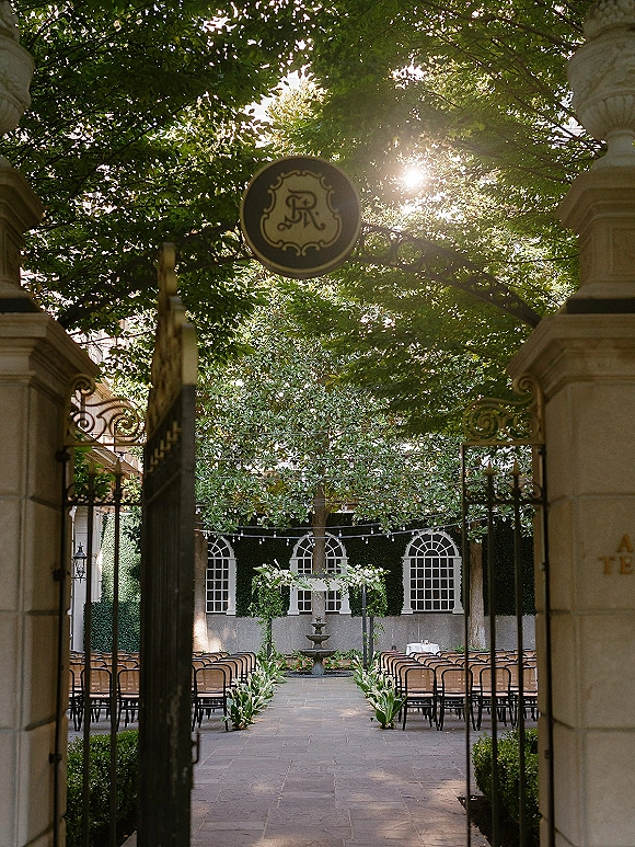 Ceremony setup with an outdoor ceremony aisle lined in greenery, facing a floral wedding arch in a sunlit courtyard with fountain and ivy wall