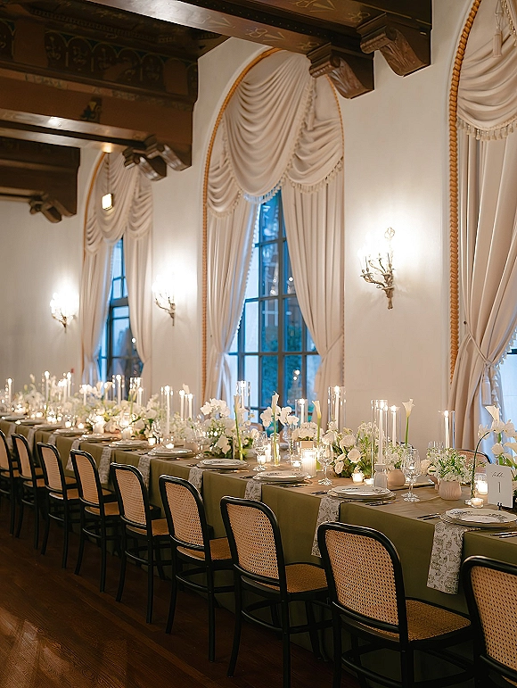 Reception tablescape with long head table decor, taper candles and white florals on olive linens, set beneath tall windows and draped curtains