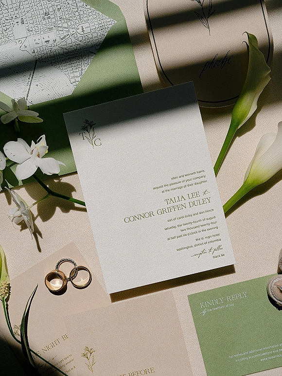 Invitation suite flat lay with wedding invitation card, RSVP, envelope, map insert, rings, calla lilies, and wax seal on linen in sun shadows