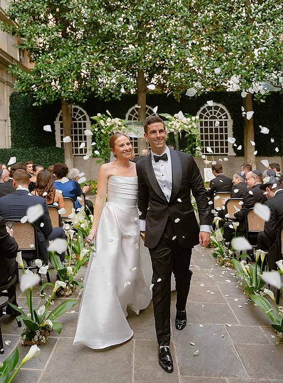 Wedding recessional with a rose petal toss as bride in strapless satin gown and groom in black tux walk a stone aisle in a courtyard