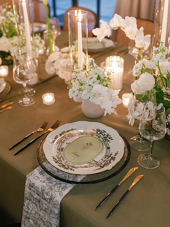 Reception tablescape wedding table setting with white orchids and roses, pillar and votive candles in glass holders, patterned plates, window light