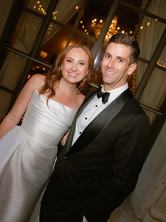 Couple portrait of bride and groom smiling in formal wedding attire, her strapless satin dress by window panes with warm string lights behind