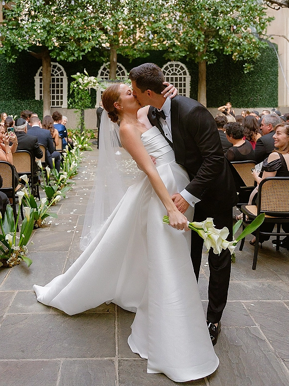 Wedding kiss portrait of a bride in strapless satin wedding dress and veil dipping with groom in tuxedo, ivy-walled courtyard aisle behind