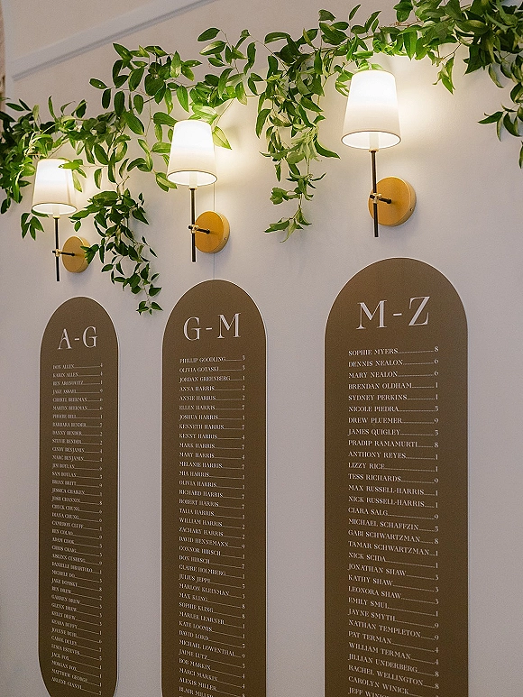 Seating chart with alphabetical guest list on arched brown boards, paired with greenery garland and wall sconces on a white wall