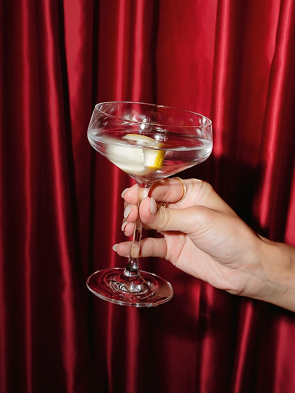 Wedding cocktail in a coupe glass with a lemon twist, held by a manicured hand with a ring against a red satin curtain backdrop