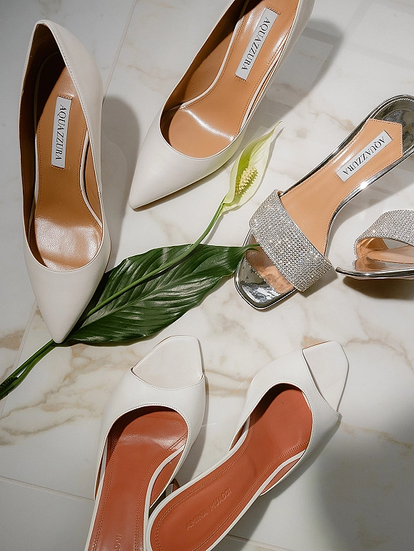 Bridal shoes flatlay featuring white wedding heels and a rhinestone slide sandal arranged on marble tile with a green leaf accent