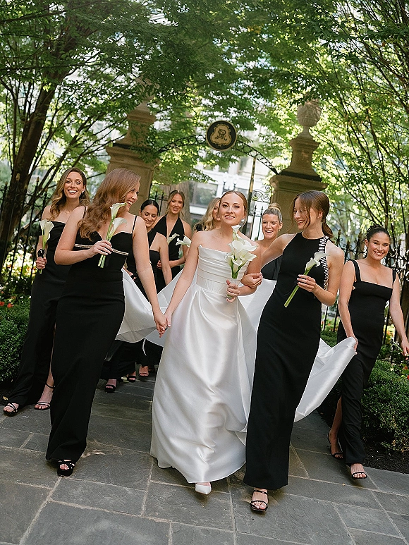 Bridesmaid group photo of the bride with bridesmaids in black dresses holding calla lily bouquets on a garden stone walkway by a wrought iron gate