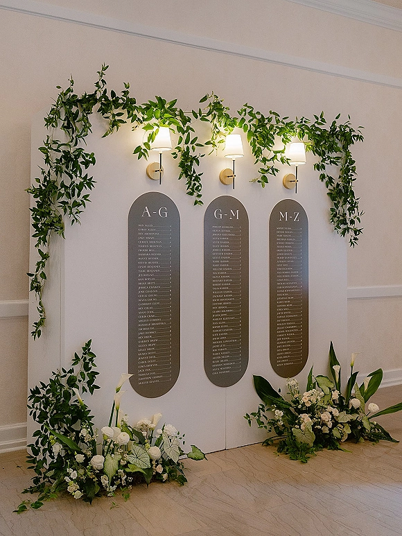 Seating chart display with wedding seating chart boards framed by greenery garland, calla lilies, and white roses against a paneled wall