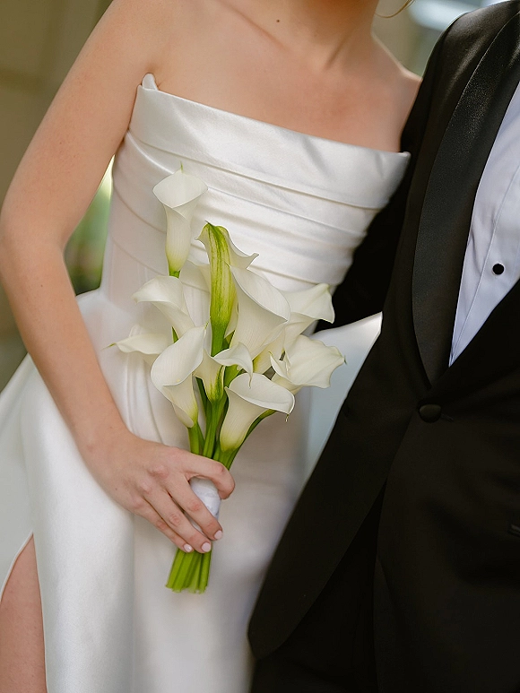 Bridal bouquet of white calla lilies with satin wrap held by bride in strapless satin dress beside groom in tuxedo, greenery blurred behind