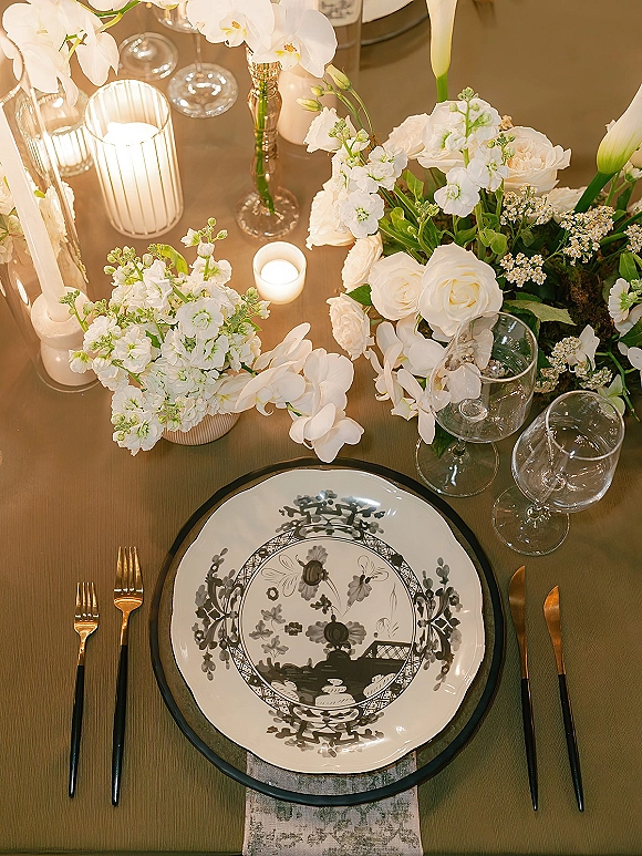 Reception tablescape wedding place setting with patterned plates, black flatware, wine glasses, and white florals in glass vases with candlelight
