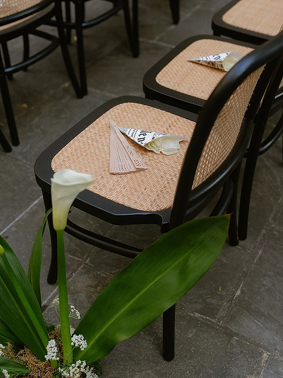 Ceremony chair decor with wedding program fan and confetti cones on black cane chairs, accented with calla lilies and greenery on a stone patio