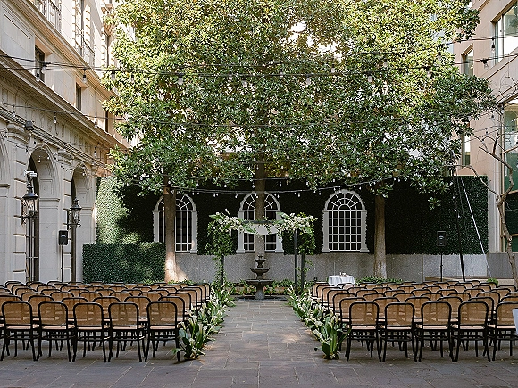 Ceremony setup with wood chairs facing a white floral arch, greenery-lined aisle, string lights overhead, and fountain in an ivy courtyard