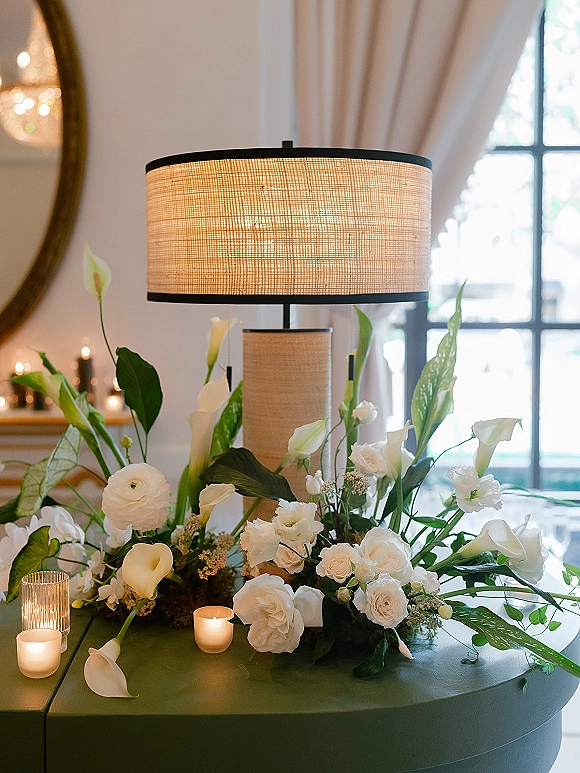 Reception centerpiece with white calla lilies and roses, greenery, bud vases, and pillar and votive candles beside a linen-shaded lamp on a round table.