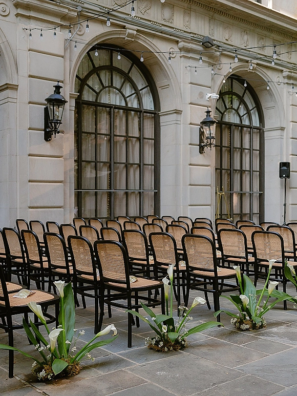 Ceremony seating with outdoor ceremony chairs in neat rows, bistro cane backs flanking calla lily aisle florals on a stone patio under string lights