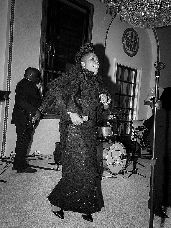 Wedding reception band with a singer in a black sequin gown holding a microphone on a ballroom stage beneath a chandelier