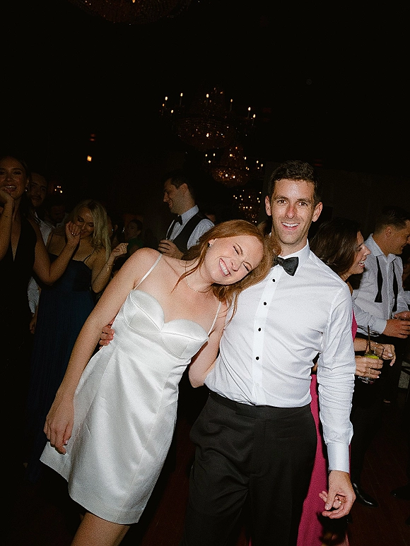 Wedding reception dancing as bride in a strapless satin dress leans on groom in bow tie, holding cocktails on a dim dance floor under chandeliers