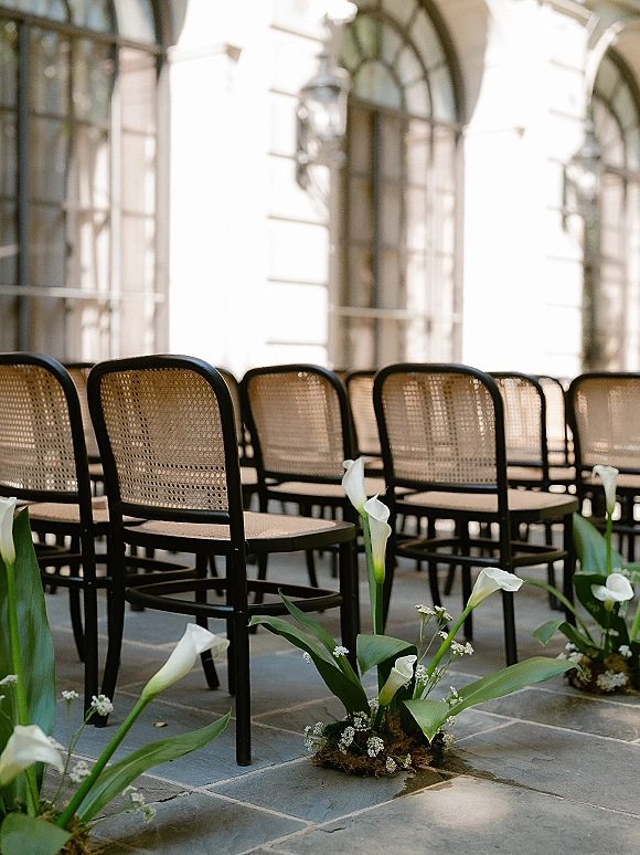 Ceremony aisle decor with white calla lily aisle flowers in moss bases, baby's breath and greenery beside cane back chairs on a stone patio