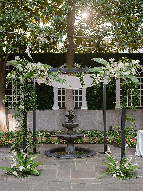 Wedding altar decor with a modern black square arch, white drapery, roses and calla lilies, greenery, and string lights in a courtyard by a fountain
