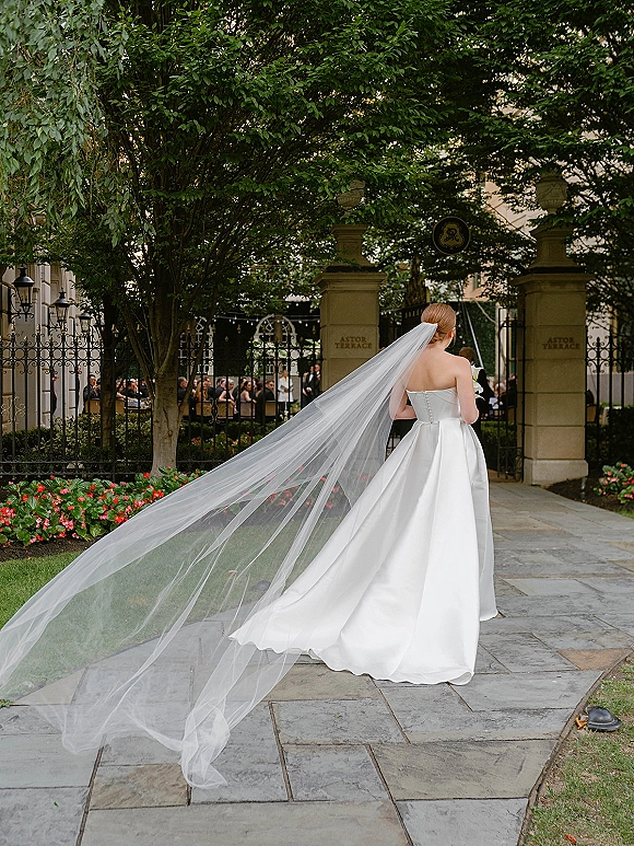 Bride portrait from behind in a strapless gown with cathedral veil train and bouquet, walking down a stone garden courtyard aisle past guests