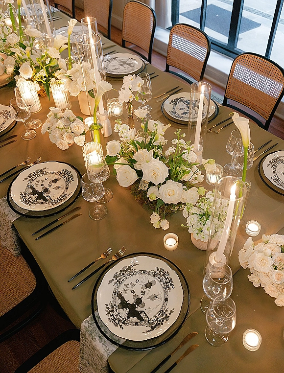 Reception tablescape with an olive green tablecloth, white floral centerpieces and candlelight, set in an indoor dining room with large windows