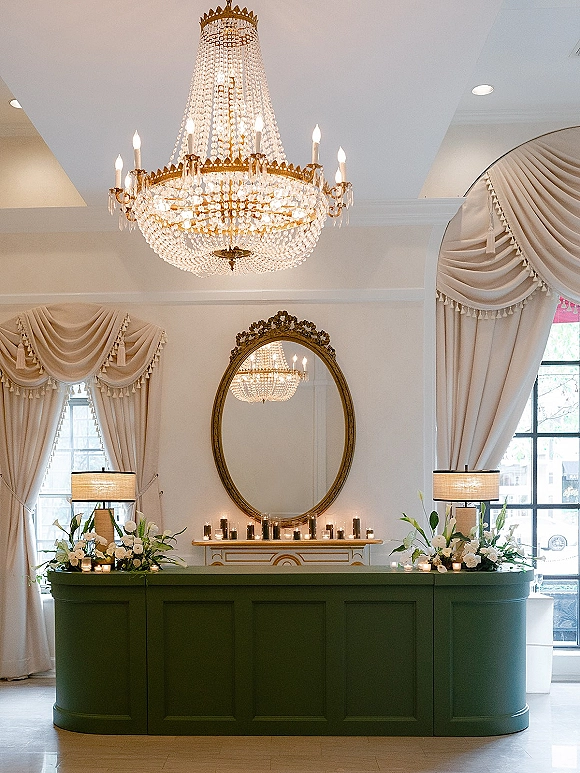 Wedding reception bar with green front, white florals and votive candles, set beneath a crystal chandelier and oval mirror in a bright lobby