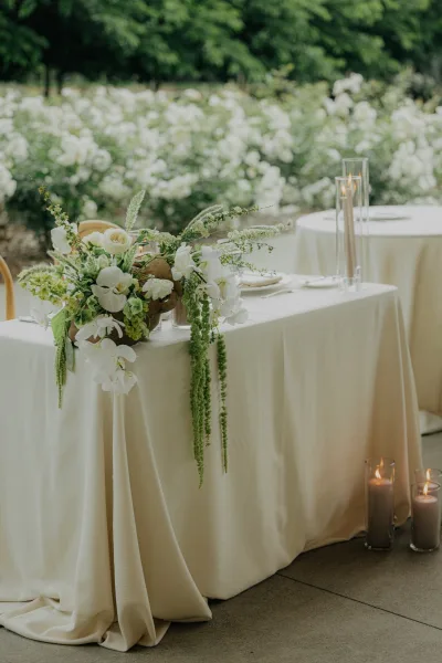 sweetheart table decor featuring a white and green centerpiece, accented by taper candles, set on an outdoor patio edged with garden bushes.