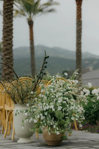 Ceremony aisle flowers styled as outdoor ceremony florals in ceramic pots, lining a stone patio framed by palm trees with mountains beyond.