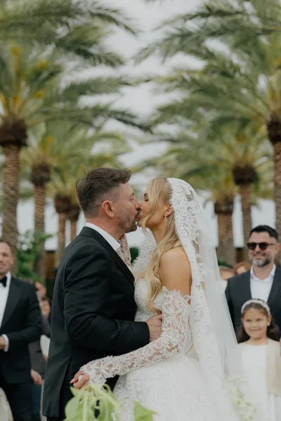 During the ceremony kiss moment, the bride in lace gown and groom in a black tuxedo embrace under palm trees and a cloudy sky.