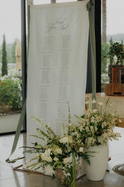 Wedding seating chart styled as a find your seat sign on a copper stand, set by tall windows overlooking garden trees and a sunlit patio.