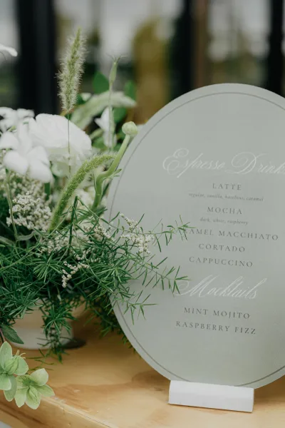 Wedding drink menu displayed as a bar menu sign, trimmed with florals on a wood surface by a glass window with blurred greenery.