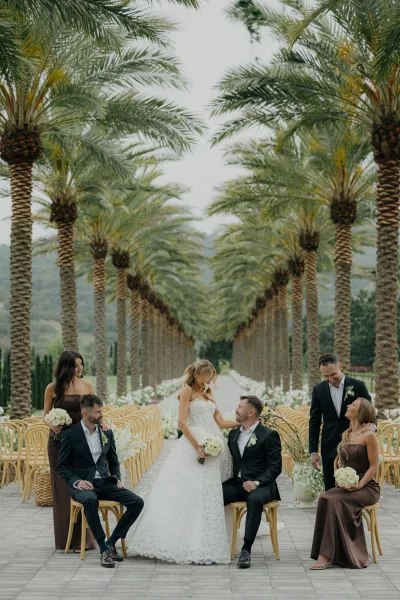 Wedding party portrait featuring bride and groom with party seated on bentwood chairs along an outdoor aisle, backed by palm trees and distant hills.