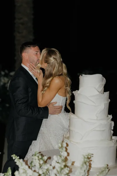 Wedding kiss portrait as the bride and groom kiss beside a ruffled white wedding cake, framed by dark greenery and a silhouetted palm at night.