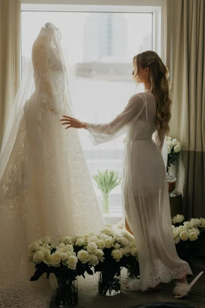 Bride getting ready with her wedding dress by a bright window, city buildings soft beyond the glass, sheer curtains framing the scene.