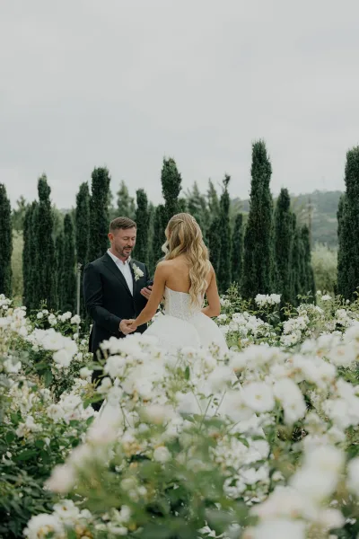 First look moment as a couple holding hands in a cypress-lined garden, framed by white garden roses with distant hills beneath an overcast sky.