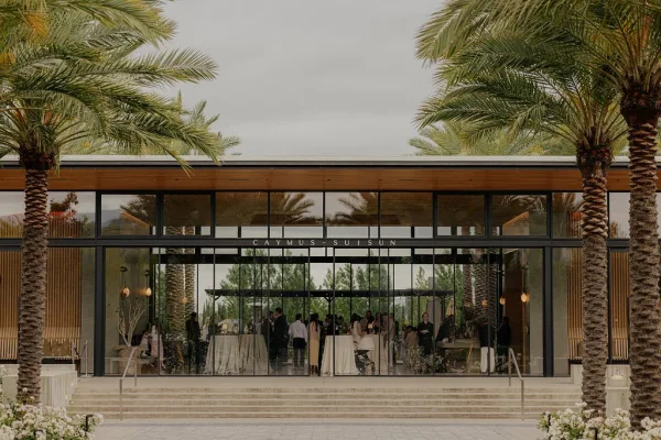 Modern wedding venue framed by palm trees and a glass facade, styled for a cocktail hour setup with floral arrangements at the entrance.