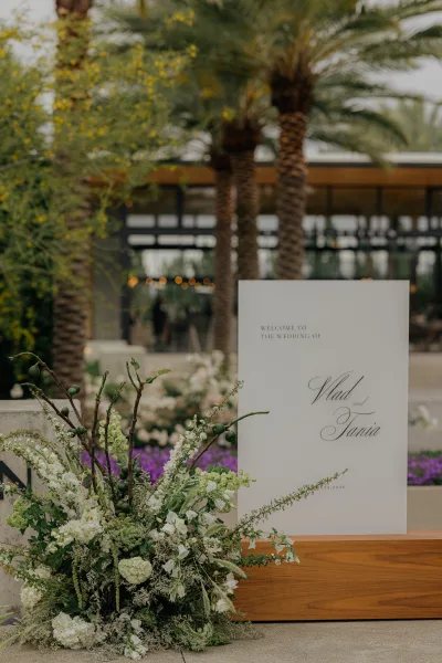 Wedding welcome sign with florals greets guests along a concrete walkway under palm trees and string lights by a modern building.