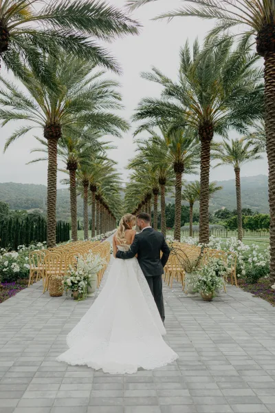 Couple portrait of a bride and groom walking away down the aisle, her ballgown trailing, with palm trees and mountains beyond.
