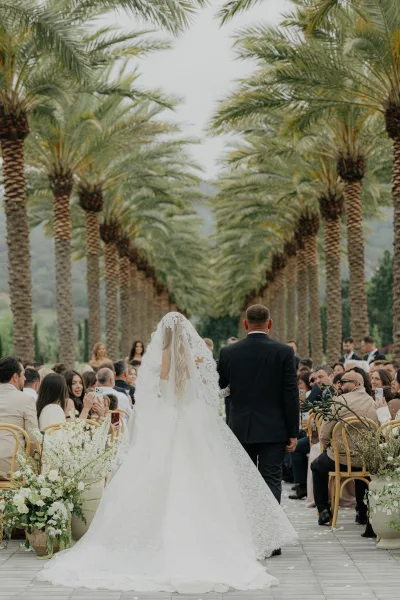Ceremony processional with a bride walking down the aisle, lace veil trailing along a stone walkway in a palm-lined outdoor garden.