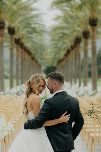 Couple portrait as the bride and groom embrace on a palm-lined garden pathway, framed by aisle florals, captured in soft natural light.