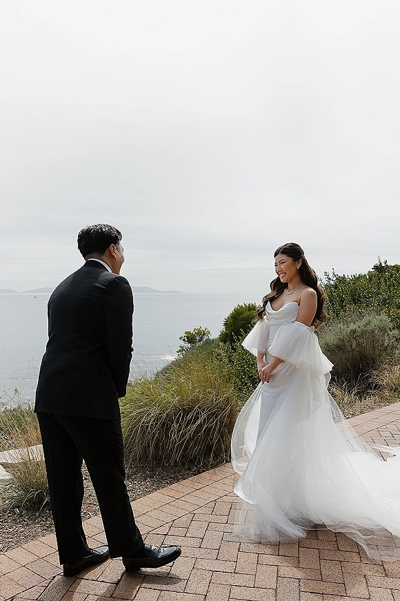 First look moment as bride in strapless tulle gown with off-shoulder sleeves meets groom in black suit on brick patio with ocean view