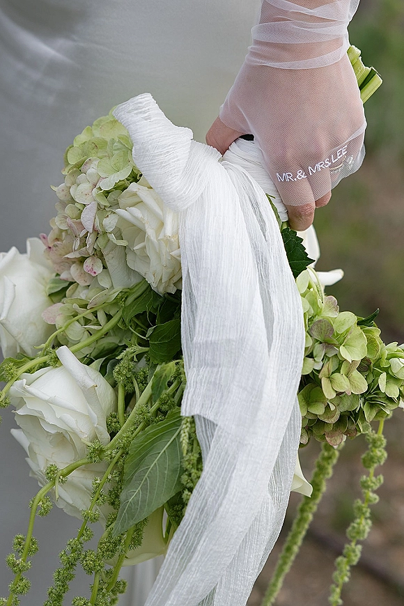 Bridal bouquet of white roses and green hydrangea with flowing ribbon, held in sheer embroidered gloves against a blurred garden path