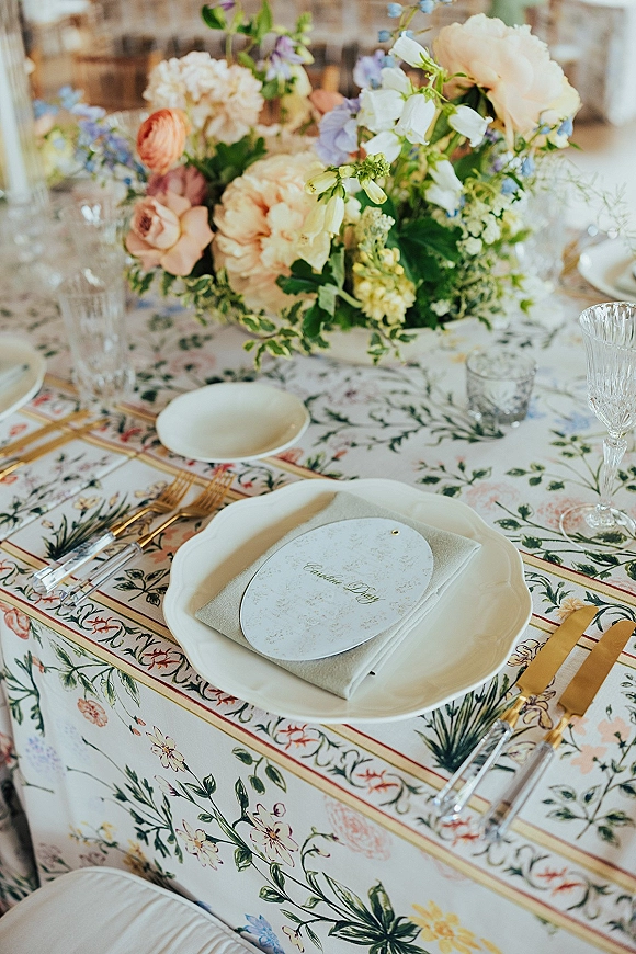 Reception tablescape with floral centerpiece, scalloped white plates, gold flatware, crystal goblets, and place cards on a patterned tablecloth