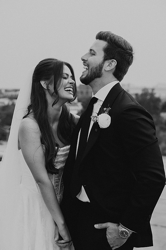 Couple portrait of bride and groom in a wedding candid laugh, she hugs him in veil and strapless dress with skyline under cloudy sky