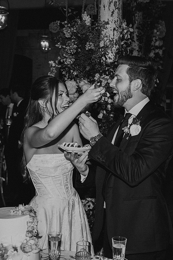 Wedding cake cutting as bride and groom feed each other bites beside a two-tier buttercream cake, in an indoor reception with hanging lights and greenery backdrop