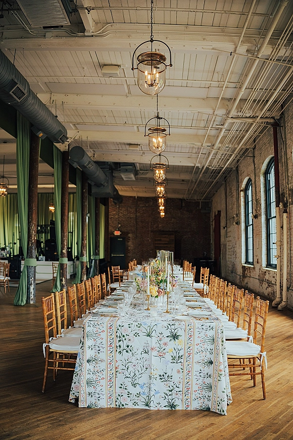Reception tablescape with a long banquet table wedding setup, botanical floral tablecloth, gold chiavari chairs, and cylinder centerpieces in a brick loft