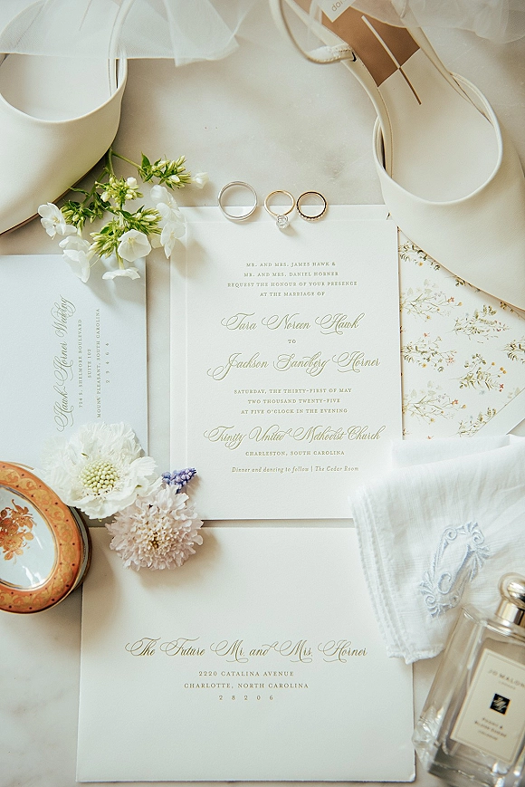 Invitation suite with classic wedding invitations, RSVP card, wax-sealed envelope, rings, white heels, perfume, and floral sprigs on a light tabletop