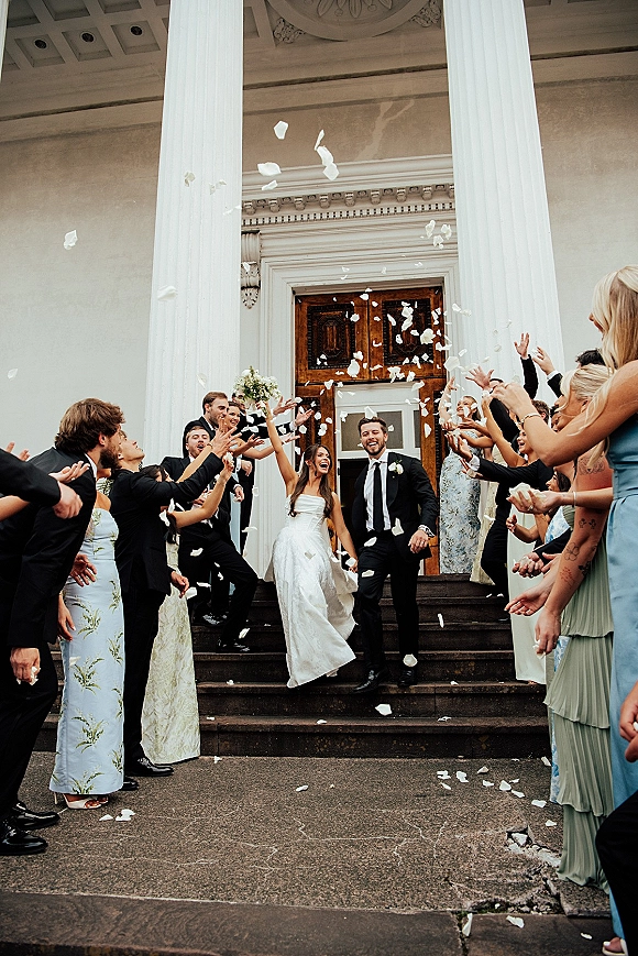 Wedding recessional as bride and groom descend stone steps while guests toss flower petals by white columns and wooden doors, bouquet raised