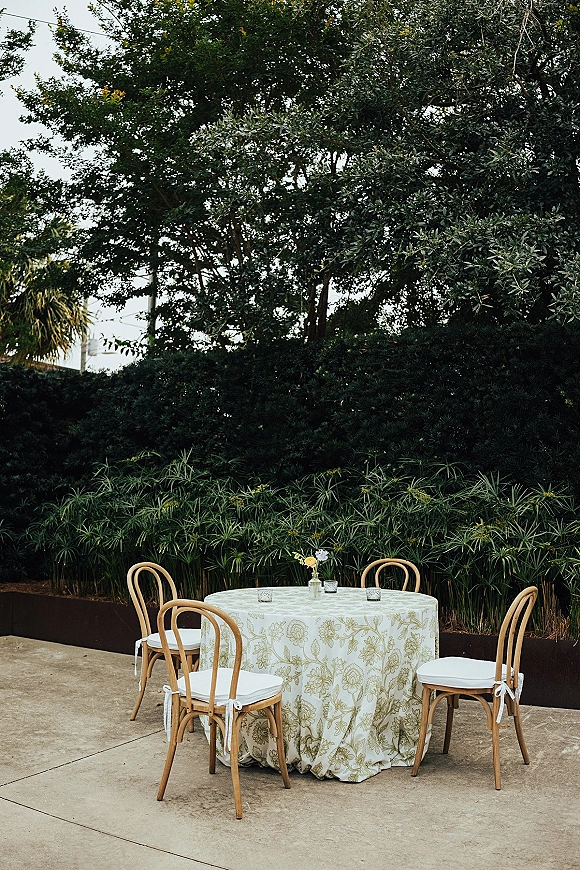 Reception tablescape with an outdoor reception table set with bud vases, small florals, and votive candles on a patio amid garden hedges
